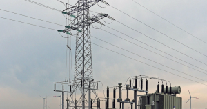 Worldwide Shortage of Electricity Transformers, image of Transformer next to a large electricity tower and wind turbines in the distance.