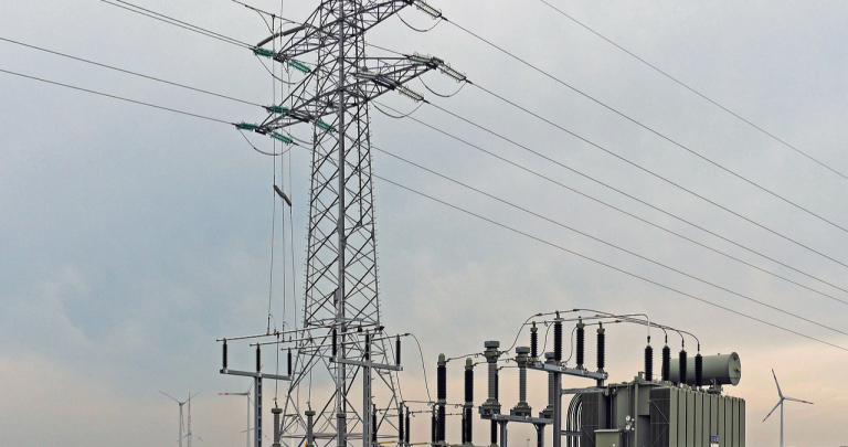 Worldwide Shortage of Electricity Transformers, image of Transformer next to a large electricity tower and wind turbines in the distance.